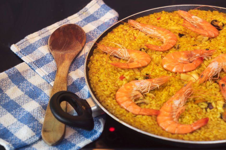 Close-up of a seafood paella with prawns in a pan, a wooden spoon, and blue towel.